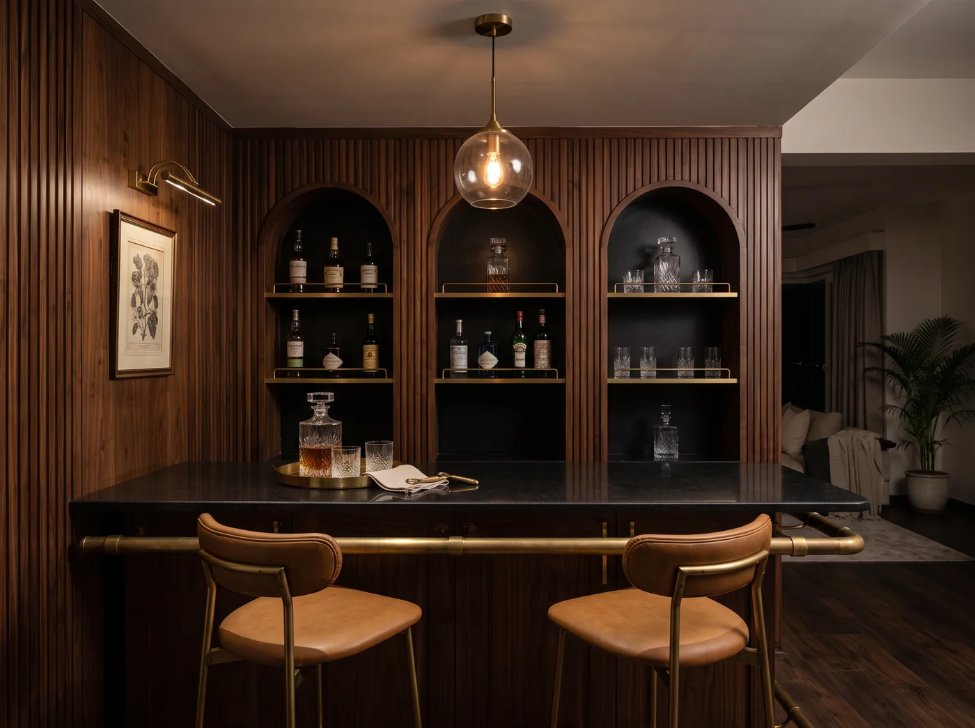 Moody home bar nook in dark fluted walnut with three arched niches lacquered black inside, walnut shelves with brass rails holding bottles and cut glass, a black granite counter over a panelled walnut bar front with a brass foot rail, a brass globe pendant, a framed botanical under a brass picture light and two tan leather stools