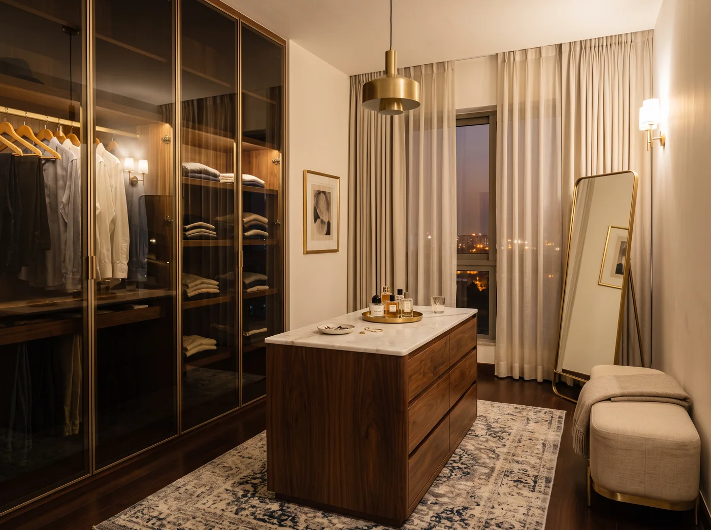Dressing room at dusk with smoked lacquered-glass wardrobes in slim brass frames lit from inside, a walnut island dresser with a white marble top under a brass drum pendant, brass wall sconces, a small framed sketch, sheer curtains at the window, a leaning brass mirror, a linen bench on brass legs and a vintage-pattern rug over a dark oak floor