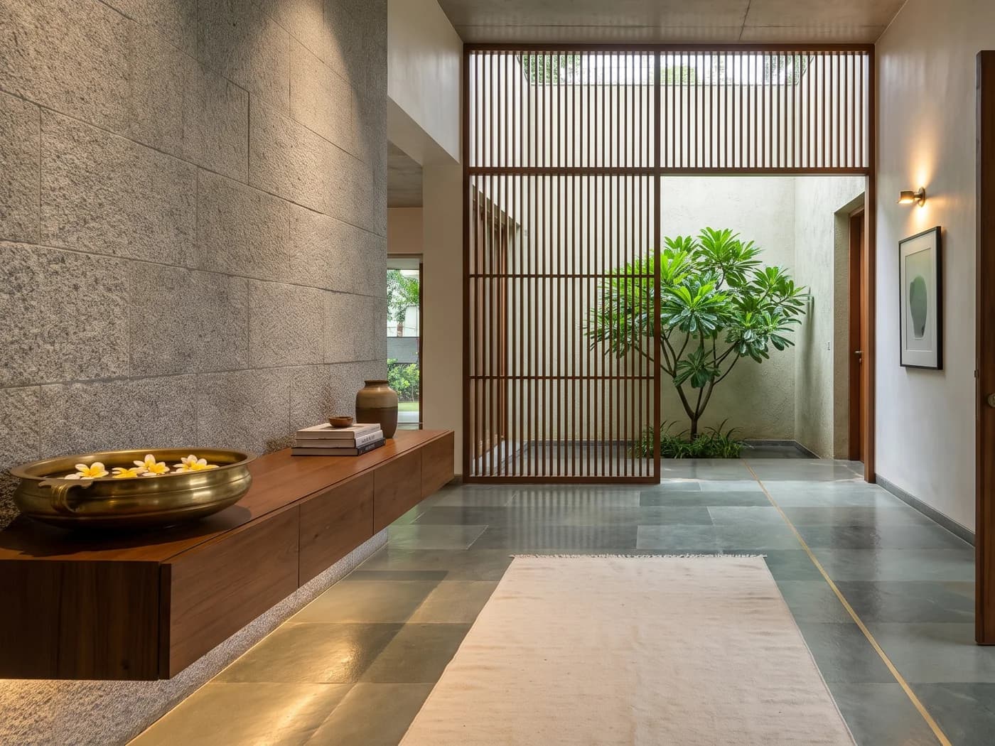 Villa entrance hall in late-afternoon monsoon light: a chiselled sadarahalli granite wall down one side, a floating Burma teak console lit from beneath carrying an unlacquered brass urli with frangipani floating in it, a stoneware jar and a stack of books, a honed kota stone floor with a brass inlay line, a full-height teak jaali onto a planted courtyard light well, and a cream cotton dhurrie running to a teak pivot door