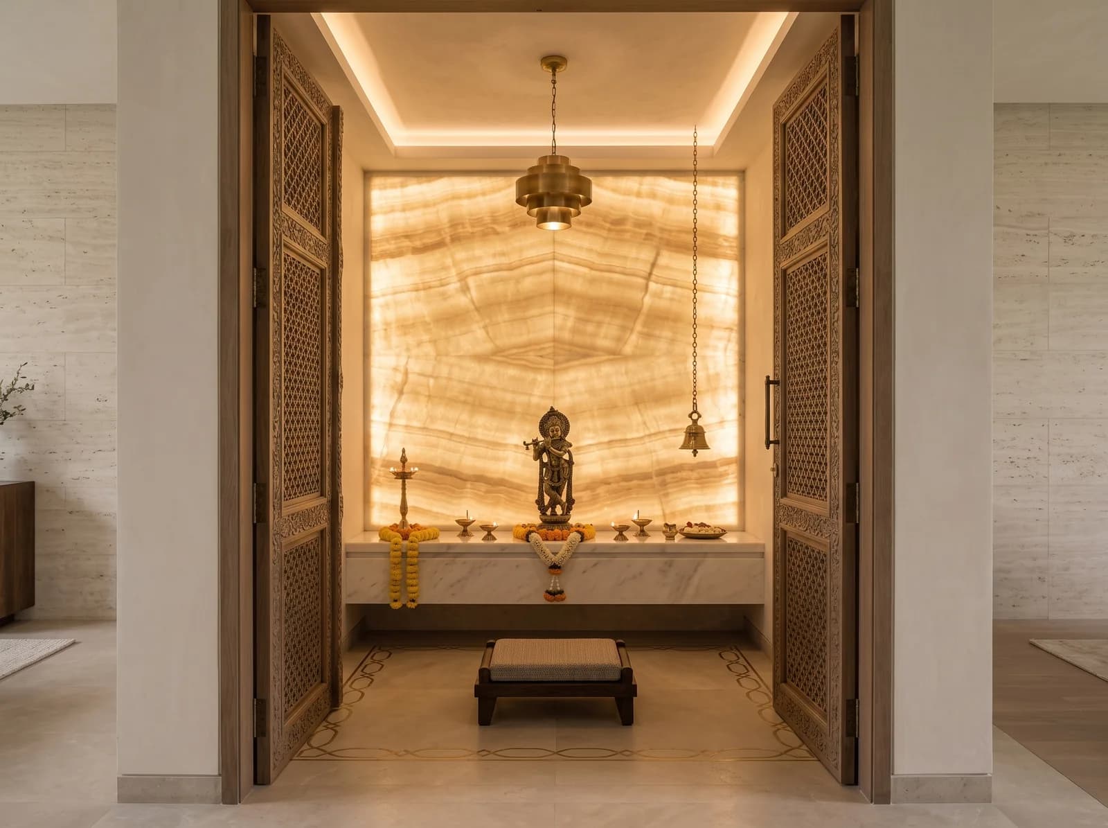 Pooja room with a backlit honey onyx wall, a marble altar with a brass idol and diyas, carved oak jaali doors, a brass pendant and marigold garlands