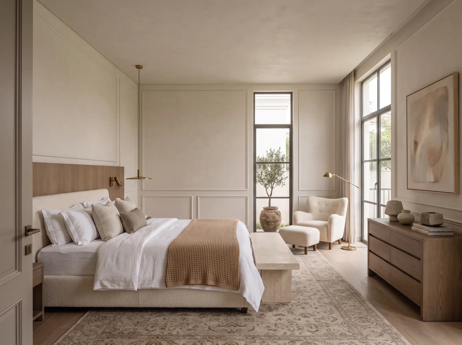 Primary bedroom with a fumed oak headboard panel, brass reading lights, a slim brass pendant, an ivory bouclé bed with a camel throw and a limestone bench