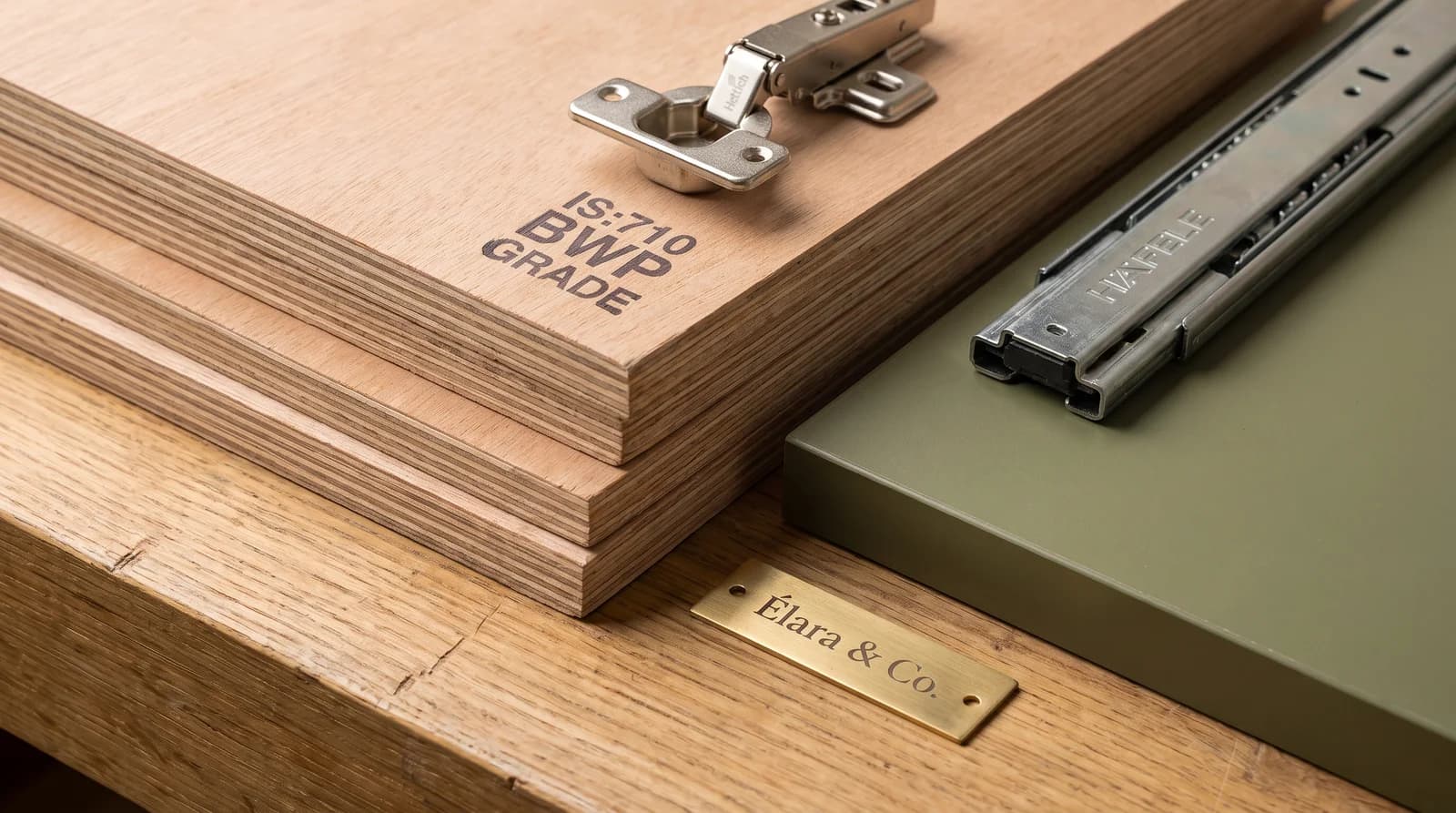 A stack of BWP-grade plywood stamped IS:710, an HDHMR board sample with an olive laminate face, and Hettich and Hafele hardware on a workshop bench